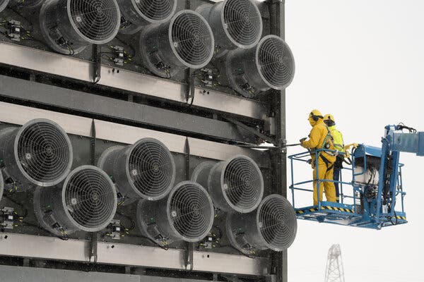 Two people in yellow work clothes and hard hats stand on a blue hydraulic lift in front of a wall covered with large, round, fan-like openings. Two people in yellow work clothes and hard hats stand on a blue hydraulic lift in front of a wall covered with large, round, fan-like openings.